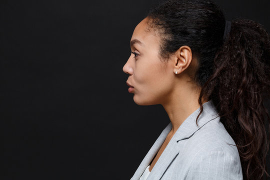 Side View Of Young African American Business Woman In Grey Suit, White Shirt Posing Isolated On Black Background Studio. Achievement Career Wealth Business Concept. Mock Up Copy Space. Looking Aside.