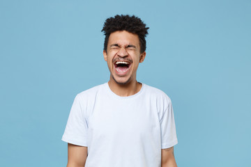 Fototapeta na wymiar Frustrated young african american guy in casual white t-shirt posing isolated on pastel blue background studio portrait. People lifestyle concept. Mock up copy space. Screaming keeping eyes closed.
