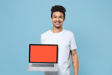 Smiling young african american guy in casual white t-shirt isolated on blue background studio portrait. People lifestyle concept. Mock up copy space. Hold laptop pc computer with blank empty screen.