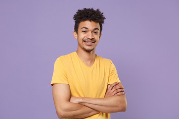 Obraz premium Smiling young african american guy in casual yellow t-shirt posing isolated on pastel violet background studio portrait. People emotions lifestyle concept. Mock up copy space. Holding hands crossed.