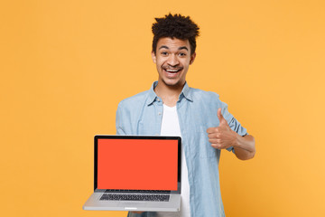 Cheerful young african american guy in casual blue shirt isolated on yellow background. People lifestyle concept. Mock up copy space. Hold laptop pc computer with blank empty screen, showing thumb up.