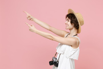 Side view smiling young tourist woman in summer dress hat with photo camera isolated on pink background. Traveling to travel weekends getaway. Air flight journey concept. Point index fingers aside up.