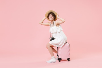 Smiling tourist girl in dress hat with photo camera isolated on pink background. Female traveling abroad to travel weekend getaway. Air flight journey concept. Sitting on suitcase put hands on head.