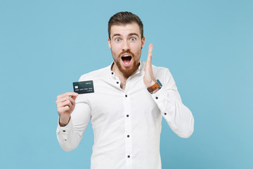 Shocked young bearded man guy 20s in white classic shirt isolated on pastel blue wall background studio portrait. People lifestyle concept. Mock up copy space. Hold credit bank card, spreading hands.