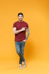 Fototapeta premium Cheerful young bearded tattooed man guy in casual t-shirt black cap posing isolated on yellow wall background studio portrait. People lifestyle concept. Mock up copy space. Point index finger aside.