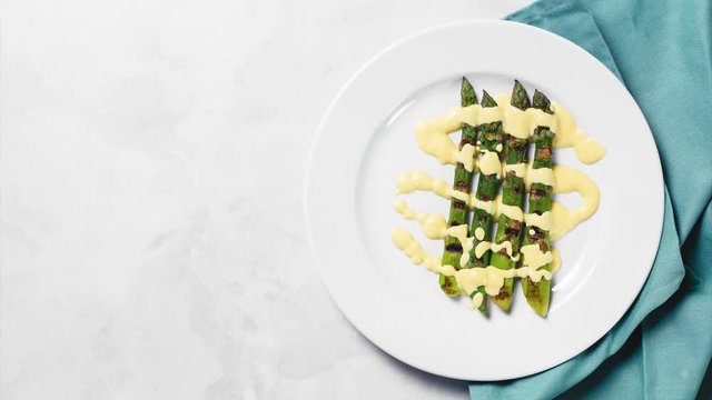 Grilled sparagus with hollandaise sauce. Restaurant background with copy space. Appearing on white palte, kitchen rag, on white concrete, top view. Stop motion animation. Mediterranean food.