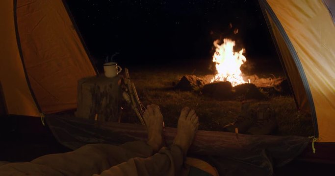 Tourist at night in a tent warms tired legs in front of a fire. Inside view. On the horizon - the starry sky.