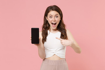Naklejka premium Excited young brunette woman girl in light casual clothes isolated on pastel pink background. People lifestyle concept. Mock up copy space. Point index finger on mobile phone with blank empty screen.