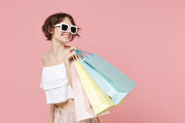 Smiling young woman girl in summer clothes, dark eyeglasses hold package bag with purchases isolated on pastel pink wall background. Shopping discount sale concept. Mock up copy space. Looking aside.