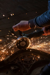 Side view of a man's hands working on a metal part of a garden bench, using an electric grinder while sparks are flying around in the industrial workshop