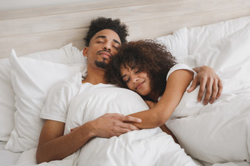 Young calm african loving couple girl guy in t-shirts lying on bed with white sheet pillow blanket...