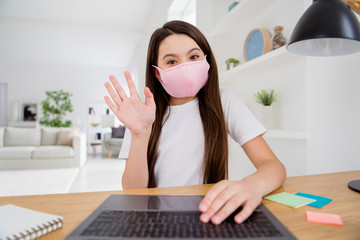 Closeup photo of little pupil school lady talk notebook online lesson video meeting teacher classmates say hi raise arm sit desk use facial mask distance quarantine study house room indoors