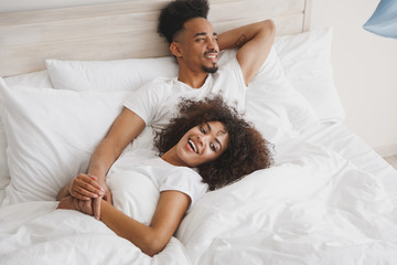 Young calm african loving couple girl guy in t-shirts lying on bed with white sheet pillow blanket indoors in bedroom at home, spending time in room. Rest relax good mood quarantine lifestyle concept.