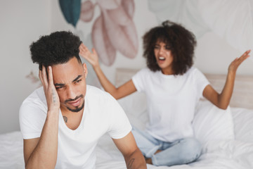 Young angry sad irritated frustrated african swearing screaming couple girl guy in white t-shirts sitting on bed indoors in bedroom at home spending time in room. Bad mood quarantine lifestyle concept