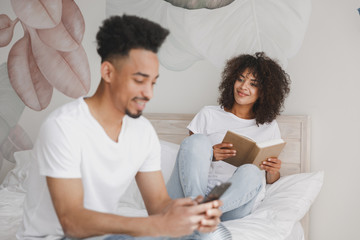 Young african loving couple girl guy in white t-shirts reading book using mobile phone lying on bed indoors in bedroom at home spending time in room. Rest relax good mood quarantine lifestyle concept.