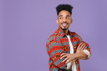 Smiling young african american guy in casual colorful shirt posing isolated on violet background studio portrait. People lifestyle concept. Mock up copy space. Holding hands crossed, looking aside.