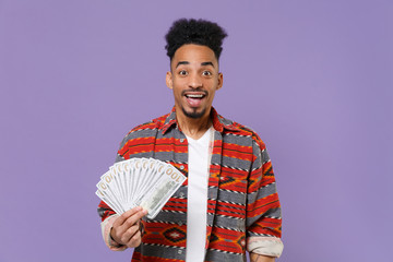 Excited young african american guy in casual colorful shirt posing isolated on violet background studio portrait. People lifestyle concept. Mock up copy space. Hold fan cash money in dollar banknotes.