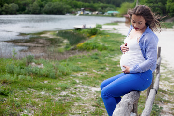 Adult pregnant woman relaxing in the nature against the lake . Pretty girl looking to the tummy  with love, female dressed blue jeans, white t-shirt, and shirt