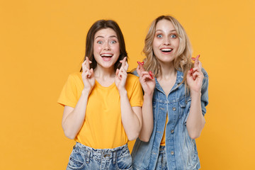 Two excited young women girls friends in casual t-shirts denim clothes posing isolated on yellow background. People lifestyle concept. Waiting for special moment, keeping fingers crossed, making wish.