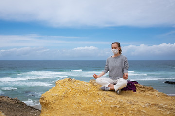 Peaceful woman in medical mask, practicing yoga on beach, meditating in lotus pose, keeping arms in zen gesture. High angle, copy space. Life balance during outbreak concept