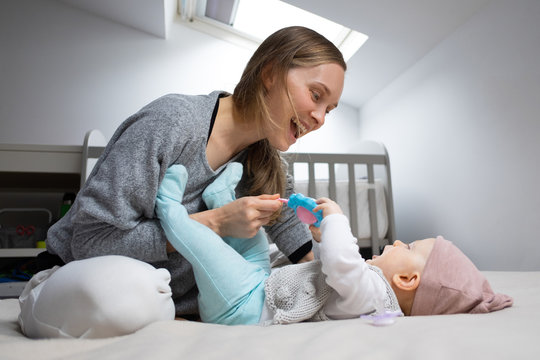Joyful mom entertaining baby with rattle toy, having fun with daughter in bedroom. Mother and little child staying at home. Child care or isolation concept