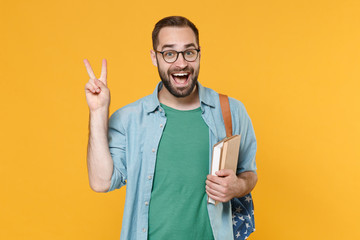 Excited young man student in casual clothes glasses with backpack hold books isolated on yellow background studio portrait. Education in high school university college concept. Showing victory sign.