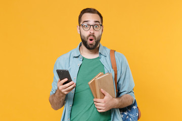Shocked young man student in casual clothes glasses with backpack hold books isolated on yellow background. Education in high school university college concept. Using mobile phone, typing sms message.