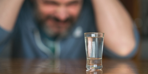 depressed man clutches his head with hands sitting at table with glass of spitrt