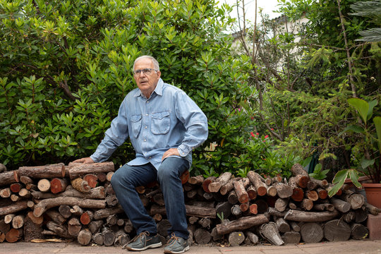 Uomo Anziano con camicia è seduto in relax nel suo giardino  sopra una ordinata atasta di legna