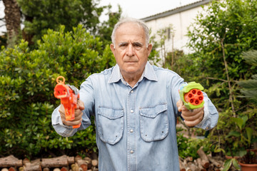 Anziano con camicia in jeans scherza allegramente nel suo giardino con due pistole ad acqua - obrazy, fototapety, plakaty