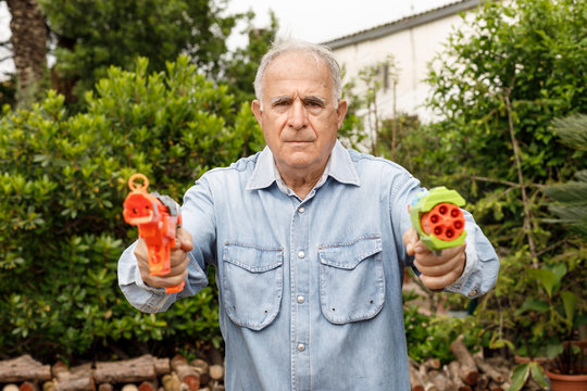 Anziano con camicia in jeans scherza allegramente nel suo giardino con due pistole ad acqua