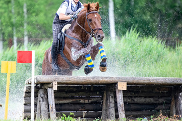 Eventing: equestrian rider jumping over an a log fence water obstacle in splash