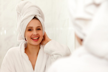 Fototapeta na wymiar Woman of Slavic appearance in dressing gown smiles in mirror. A happy lady looks in bathroom mirror, touching healthy skin of her face. Gentle and cheerful girl after shower at mirror