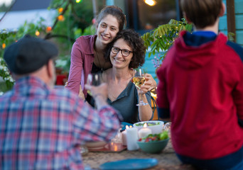 in the evening, dinner with the family around a table in the garden, in front of the wooden house. Everyone is having fun while a young woman gives her mother a hug.