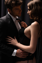seductive woman pulling tie of handsome man in suit on black