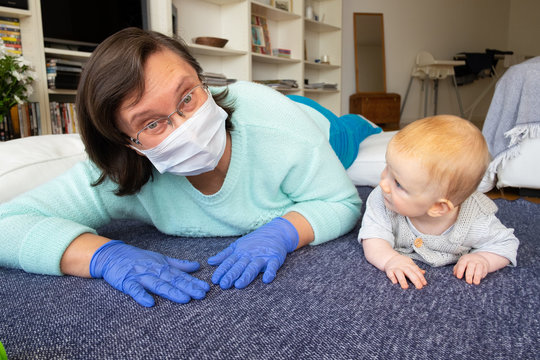 Cheerful Babysitter In Mask And Gloves Training Baby To Crawl, Playing With Child On Floor At Home. Closeup Shot. Quarantine Or Babysitting Concept