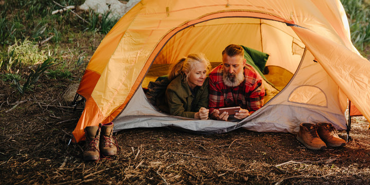 Caucasian elderly couple browsing on phone while laying in bedroll in tent