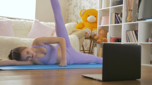 A teenage girl is engaged in fitness at home in the living room, watching the video tutorials at the laptop screen.