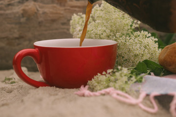 
A red cup in which coffee is poured from a Turk, in the background are white wildflowers