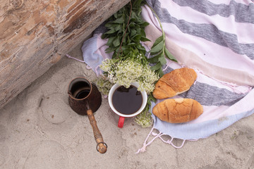 Picnic on the sea: on a beach towel are a copper Turk, a cup of coffee, croissants, white wildflowers