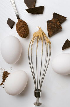 Vertical image.Electric whisk with chocolate cream on it, eggs, chocolate, cocoa powder on the white table.Concept of preparing delicious cream for confections