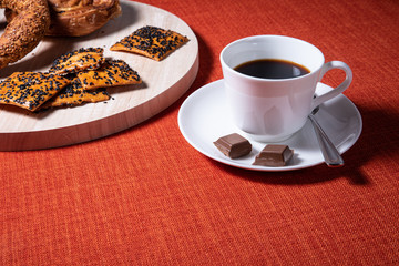 Cup of coffee with chocolate on the side. Crispy brown baked biscuits with black sesame seeds on wooden board. Bright red textile backdrop for copy space.Restaurant of cafe bites for tea, coffee
