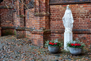 Vilnius, Lithuania - Church of St. Anne, a white statue of the Virgin Mary in the courtyard in the Catholic Church against the backdrop of a brick wall, next to paving pots with red flowers