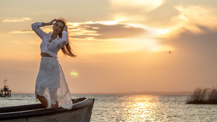 Fototapeta premium A girl in a light white dress stands in a boat and basks in the sunset. Text space.