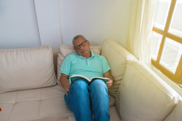 An old retired man reading a book on his couch by the window