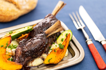 Glazed braised lamb shank on potato puree with large pieces of pumpkin and zucchini on a silver tray on a blue tablecloth aside vintage silver serving utensils and bread loaf.