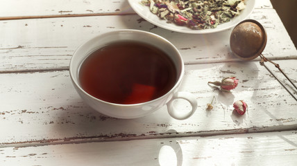Herbal tea - lemon balm, linden, verbena, rosebuds, lavender in a white plate and in a white cup, and tea infuser spoon on a light wooden table
