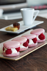 On a wooden table, four pink raspberry macaroons are arranged on a rectangular plate in front of another plate in which is arranged a coffee cup with a canelé and a financier.