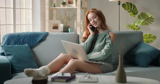 Emotional caucasian redhead high school student is talking on phone with her friend while browsing social network on laptop, positively smiling - communication concept 4k footage