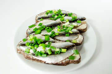 three delicious sandwiches with rye bread, butter, small sardines and green onions on a white plate on a white background